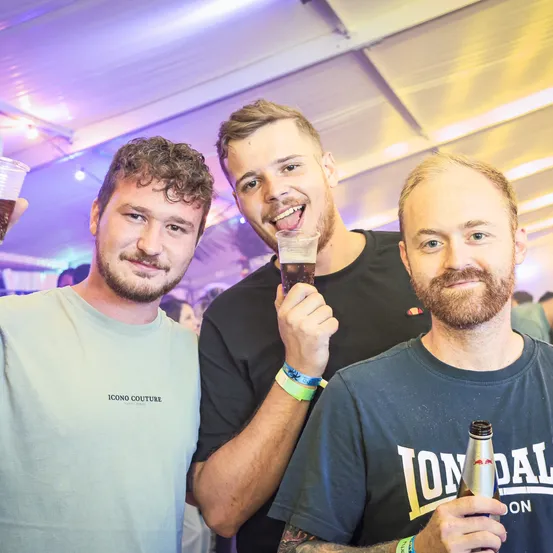 Drei Männer stehen zusammen auf einer Party. Der Mann auf der linken Seite hat ein Glas Bier in der Hand. Der Mann in der Mitte hält ein Glas Bier und lächelt. Der Mann auf der rechten Seite hält eine Flasche Bier.