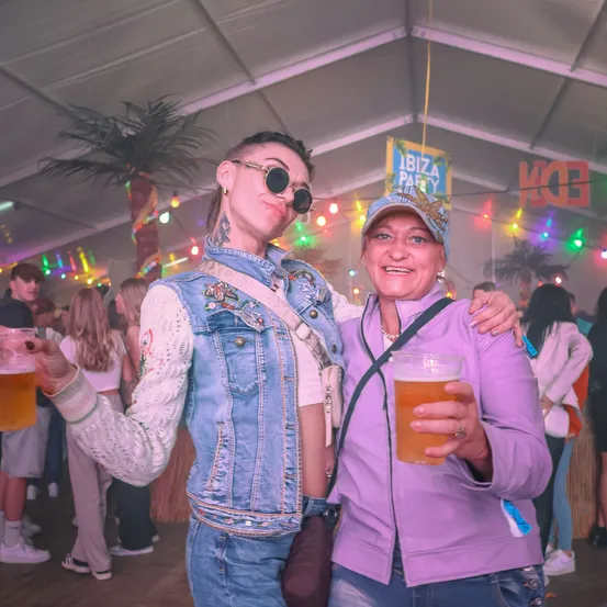 Zwei Frauen halten Getränke in der Hand, lächeln unter einem Zelt mit einem Banner, das 'Ibiza Party' sagt, und bunten Lichtern. Sie sind von einer lebhaften Menge umgeben.
