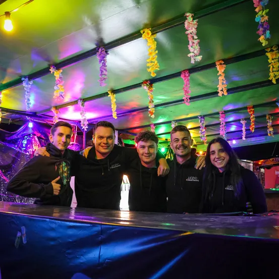 Fünf Personen stehen lächelnd hinter einer Bar und tragen passende schwarze Kapuzenpullover. Die Decke ist mit bunten Papiergirlanden und Lichtern geschmückt.