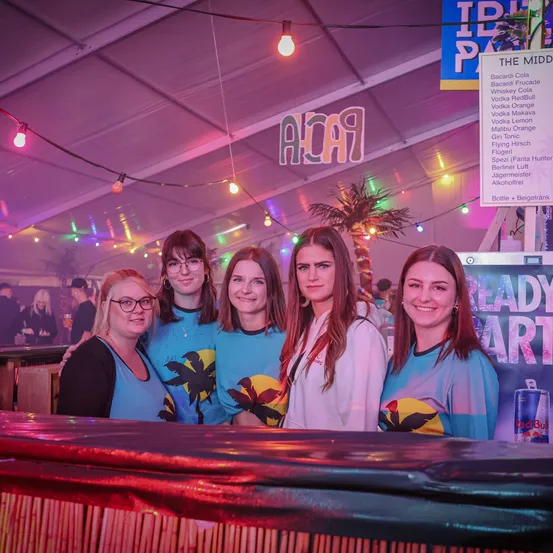 Fünf Frauen in blauen Shirts mit Palmenmotiven stehen lächelnd hinter einer Bar vor der Kamera. Dahinter zeigt ein bunt beleuchtetes Zelt eine Getränkekarte an.