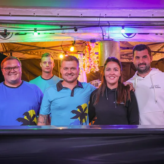 Fünf Menschen stehen in einer Bar. Eine Frau steht in der Mitte, und zwei Männer stehen auf jeder Seite. Dahinter hängen bunte Lichter an der Decke.