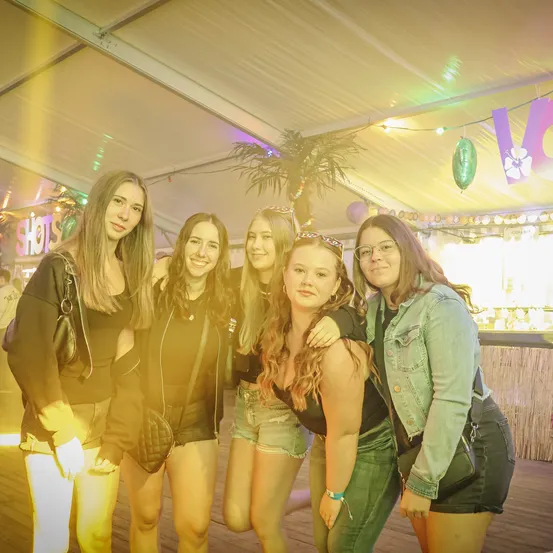Fünf junge Frauen posieren für ein Foto in einem Partyzelt mit festlichen Dekorationen, Luftballons und einem Schild mit der Aufschrift 'Shots.'