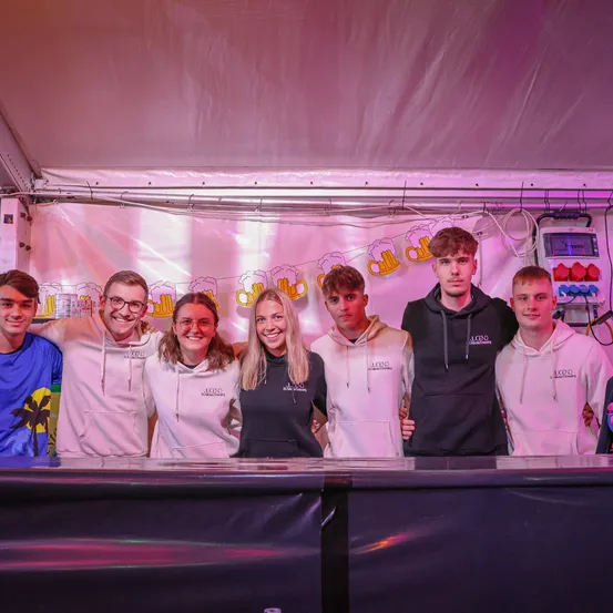 Eine Gruppe junger Menschen steht hinter einer Bar, lächelt und posiert für ein Foto. Sie tragen verschiedenfarbige Hoodies und Brillen. Ein Banner mit Bierkrügen ist im Hintergrund.