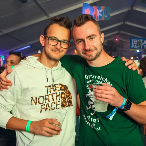 Zwei Männer lächeln und posieren für ein Foto auf einer Party, Getränke in den Händen. Einer trägt ein grünes Shirt mit Grafikdesign, der andere eine weiße Kapuzenjacke mit 'The North Face'. Hinter ihnen eine farbenfrohe Partyatmosphäre mit hängenden Schildern.
