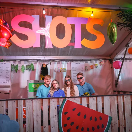 Vier Personen, zwei Männer und zwei Frauen, stehen hinter einem Tresen mit einem Wassermelonen-Ausschnitt, unter einem Neonschild, das 'SHOTS' steht. Dahinter zeigt ein Menübrett die Preise an.