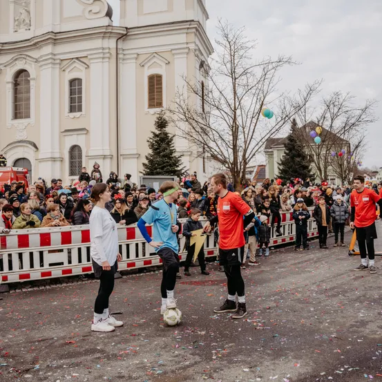 Drei Personen auf der Straße, zwei spielen Fußball, während eine Menschenmenge hinter einer Barriere zuschaut. Ein Gebäude mit Bäumen im Hintergrund.