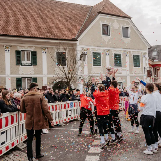 Eine Gruppe von Menschen in roten Trikots und Turnschuhen feiert auf einer Straße mit Konfetti. Hinter ihnen beobachtet eine Menge von einer Barriere aus. Im Hintergrund ein Gebäude mit braunem Dach und grünen Fensterläden.