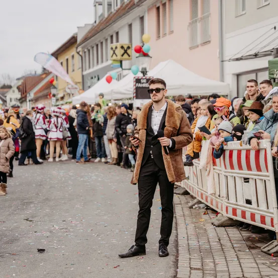 Ein Mann in einem Pelzmantel steht in der Mitte einer Straße und sieht einer Menge mit Luftballons und Zelten im Hintergrund zu.