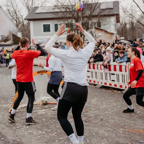 Eine Gruppe von Menschen in Sportbekleidung läuft auf einer Straße, während eine Menge zuschaut. Eine Frau in einem weißen Sweatshirt hebt ihre Hände. Hinter ihnen trennt eine rote und weiße Absperrung die Läufer von den Zuschauern.