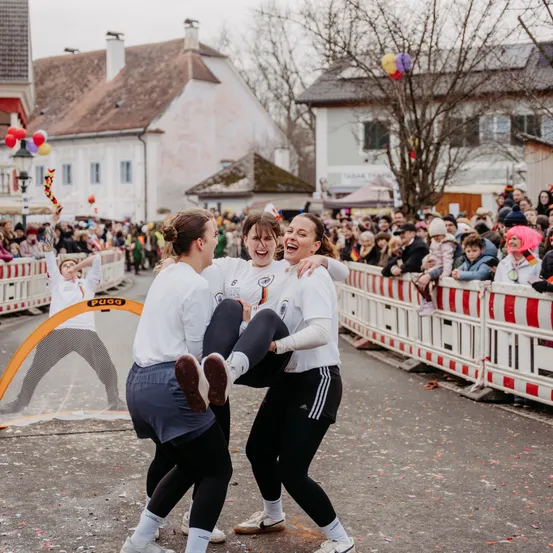 Drei Frauen in Sportkleidung kämpfen spielerisch auf der Straße, während eine Menge hinter Barrieren zusieht. Die Szene beinhaltet bunte Luftballons und festliche Dekorationen.