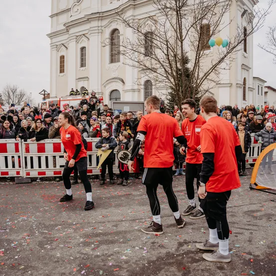 Eine Gruppe von Menschen in roten Trikots feiert auf der Straße vor einer Kirche, während eine Menge zuschaut. Einige halten Flaggen, und bunte Konfetti liegen auf dem Boden.