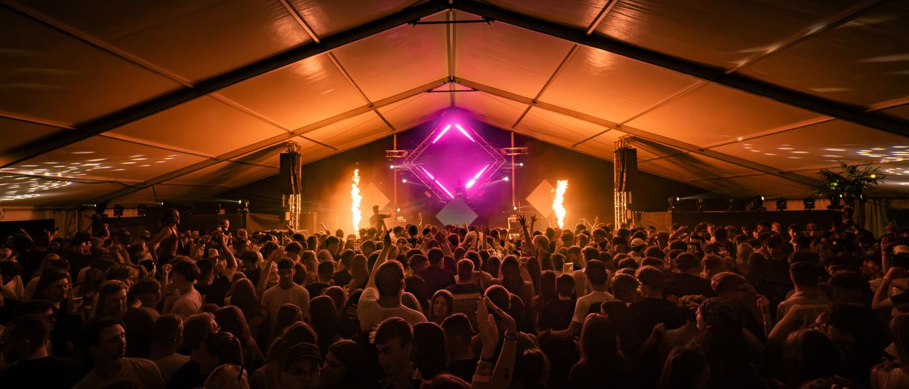 Eine große Menschenmenge versammelt sich in einem Zelt für ein Konzert, mit einer Bühne in der Mitte, die von einem lila Licht beleuchtet wird.