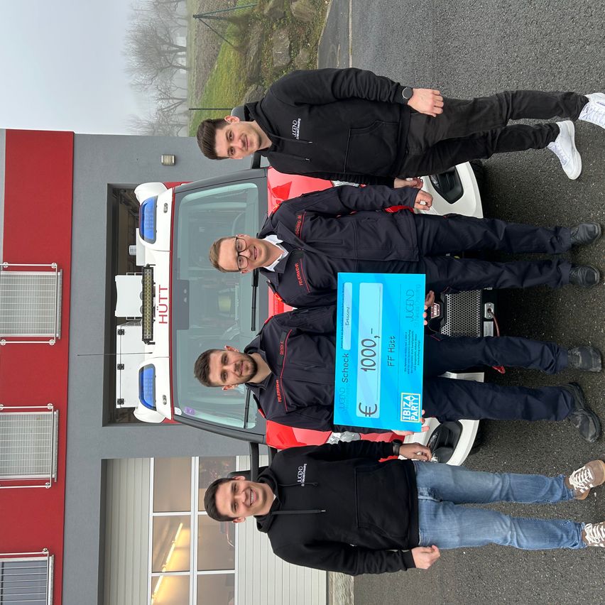 Fünf Männer stehen hinter einem roten Auto und halten einen blauen Scheck über 1000 Euro. Sie sind in schwarzen Jacken und Hosen gekleidet, einer trägt eine Brille. Sie posieren für ein Foto.