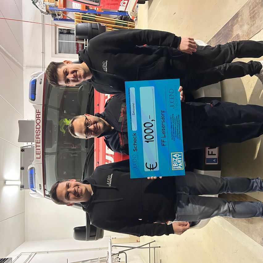 Drei Männer stehen lächelnd vor einem Feuerwehrauto. Einer hält einen blauen Scheck über 1000 Euro mit einem Partylogo.