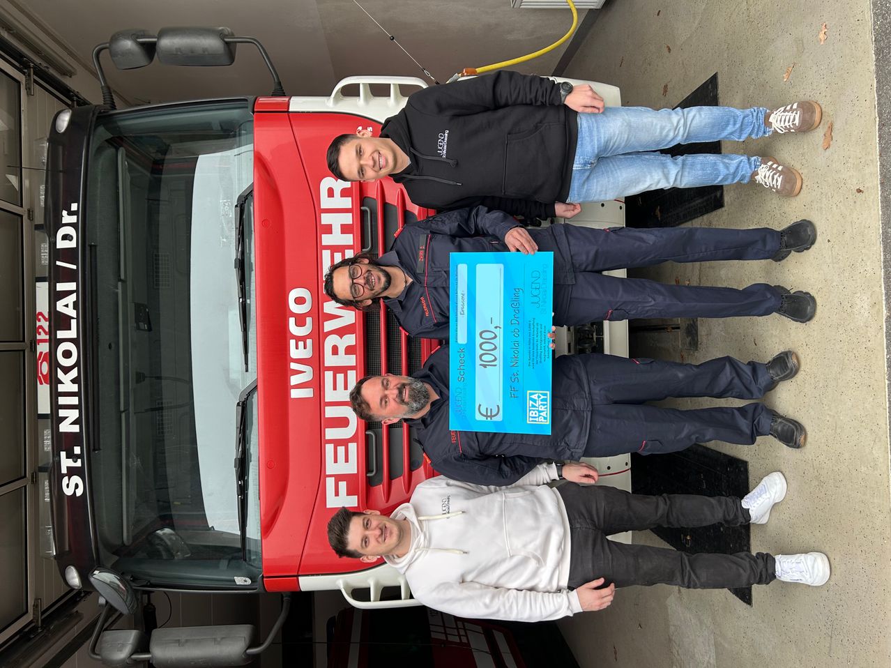 Vier Männer posieren vor einem roten LKW mit 'IVECO' und 'FEUERWEHR'. Sie halten einen blauen Gutschein für 1000 Euro.
