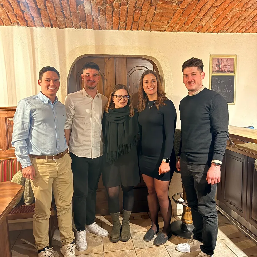 Fünf Personen stehen in einem Restaurant zusammen. Sie lächeln und scheinen für ein Foto zu posieren. Hinter ihnen befindet sich eine große Holztür und eine Wand mit einem gerahmten Bild.