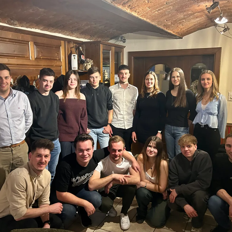 Eine Gruppe junger Erwachsener posiert für ein Foto in einem Raum mit Holzwänden und einer Ziegeldecke. Sie lächeln und einige hockten.