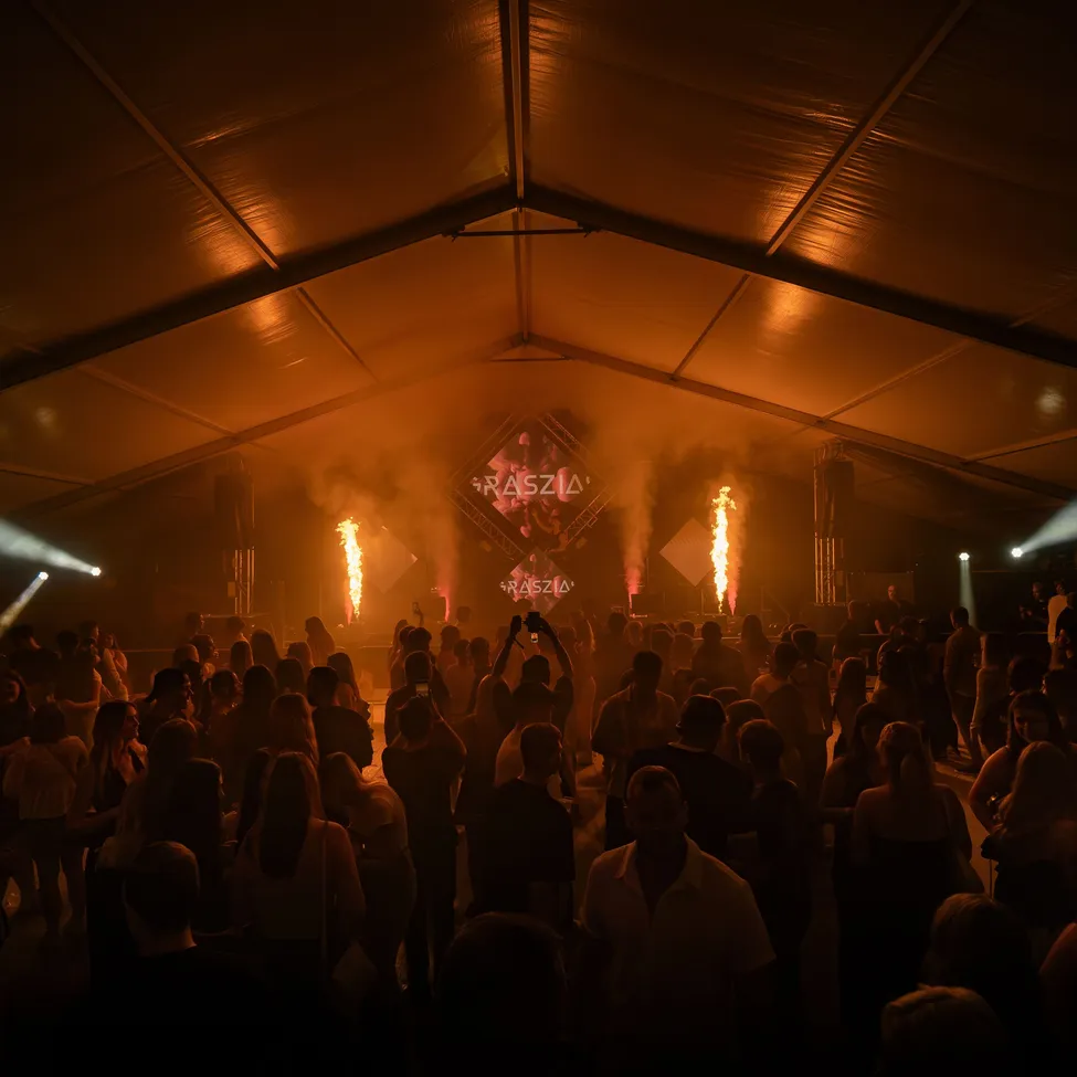 Eine große Menschenmenge versammelt sich in einem Zelt für ein Konzert. Die Bühne ist mit roten Lichtern beleuchtet und es gibt Feuereffekte.