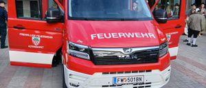 Ein roter MAN-Feuerwehrwagen mit dem Kennzeichen FW 501BN vor einem Gebäude geparkt. Personen in traditioneller Kleidung stehen in der Nähe.