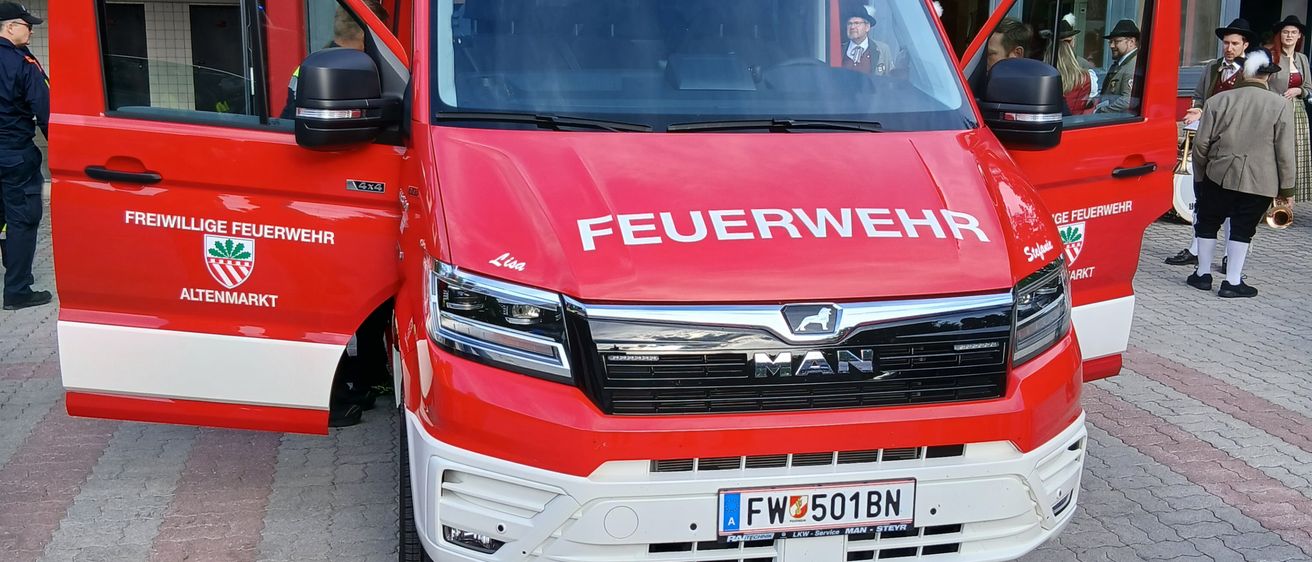 Ein roter MAN-Feuerwehrwagen mit dem Kennzeichen FW 501BN vor einem Gebäude geparkt. Personen in traditioneller Kleidung stehen in der Nähe.