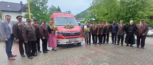 Eine Gruppe uniformierter Beamter und Zivilisten steht um einen roten Feuerwehrwagen und überreicht Blumen vor einem Bergpanorama.