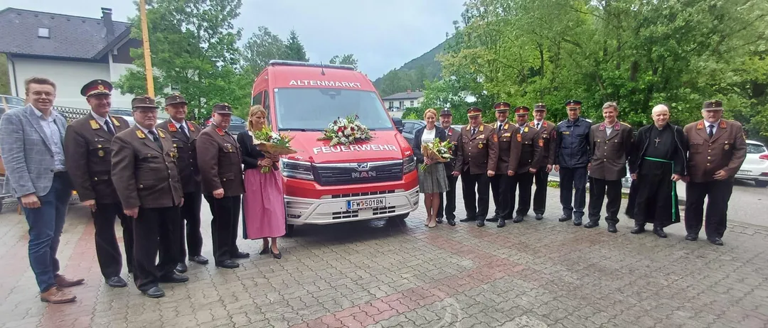 Eine Gruppe uniformierter Beamter und Zivilisten steht um einen roten Feuerwehrwagen und überreicht Blumen vor einem Bergpanorama.