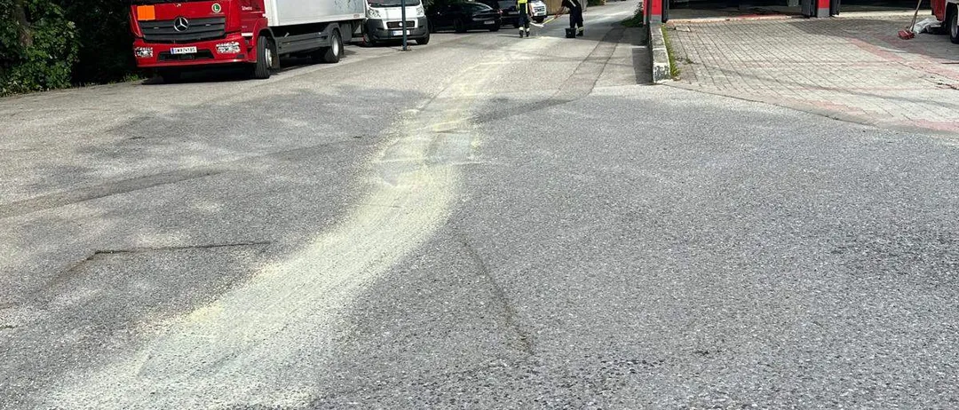 Ein roter und weißer Lkw ist am Straßenrand geparkt, in der Nähe einer Feuerwache. Bäume und geparkte Autos sind im Hintergrund.