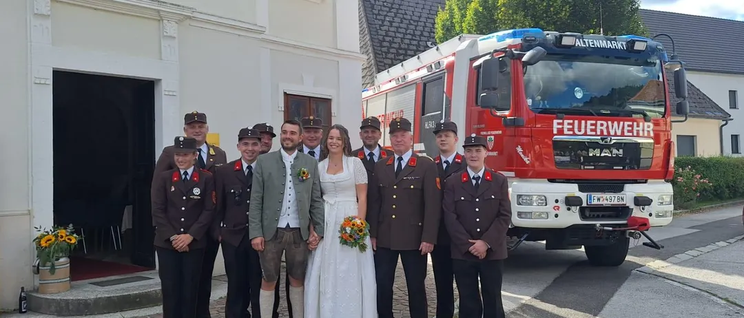 Eine Hochzeitsgesellschaft steht vor einer Kirche mit einem Feuerwehrauto im Hintergrund.