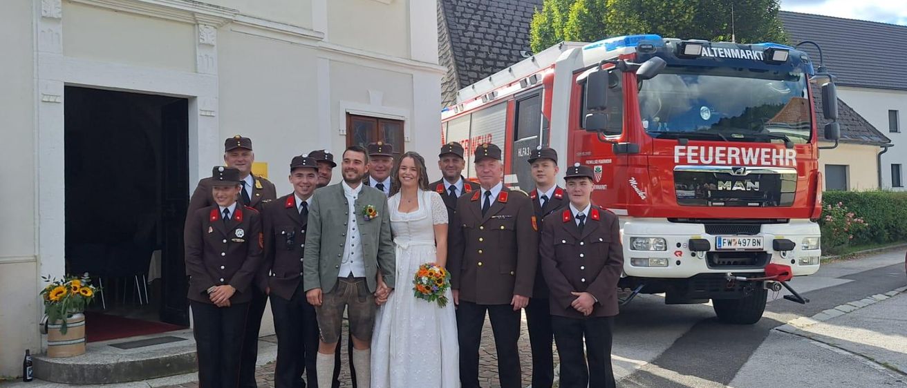 Eine Hochzeitsgesellschaft steht vor einer Kirche mit einem Feuerwehrauto im Hintergrund.