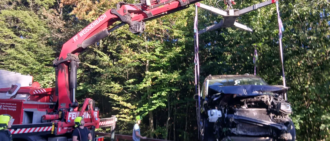 Ein roter Kran hebt ein beschädigtes Auto vom Boden, mit einer Person in einem gelben Helm in der Nähe. Eine andere Person beobachtet vom Straßenrand aus.
