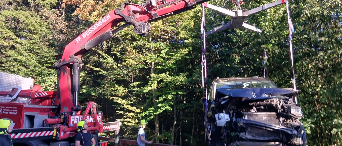 Ein roter Kran hebt ein beschädigtes Auto vom Boden, mit einer Person in einem gelben Helm in der Nähe. Eine andere Person beobachtet vom Straßenrand aus.