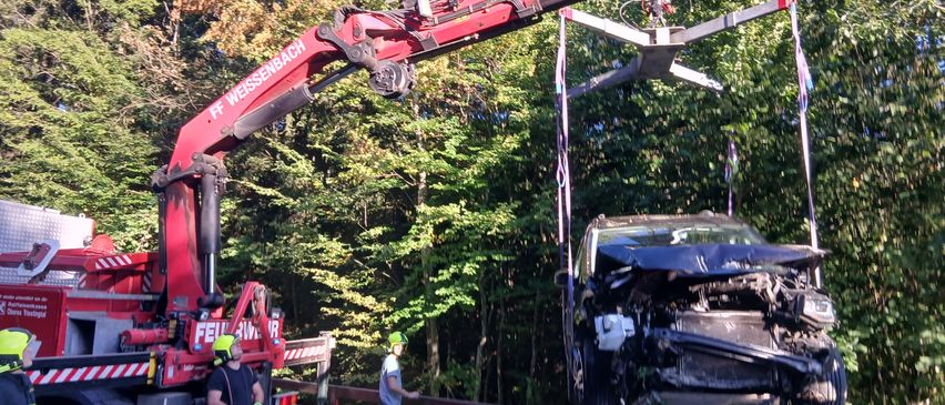 Ein roter Kran hebt ein beschädigtes Auto vom Boden, mit einer Person in einem gelben Helm in der Nähe. Eine andere Person beobachtet vom Straßenrand aus.