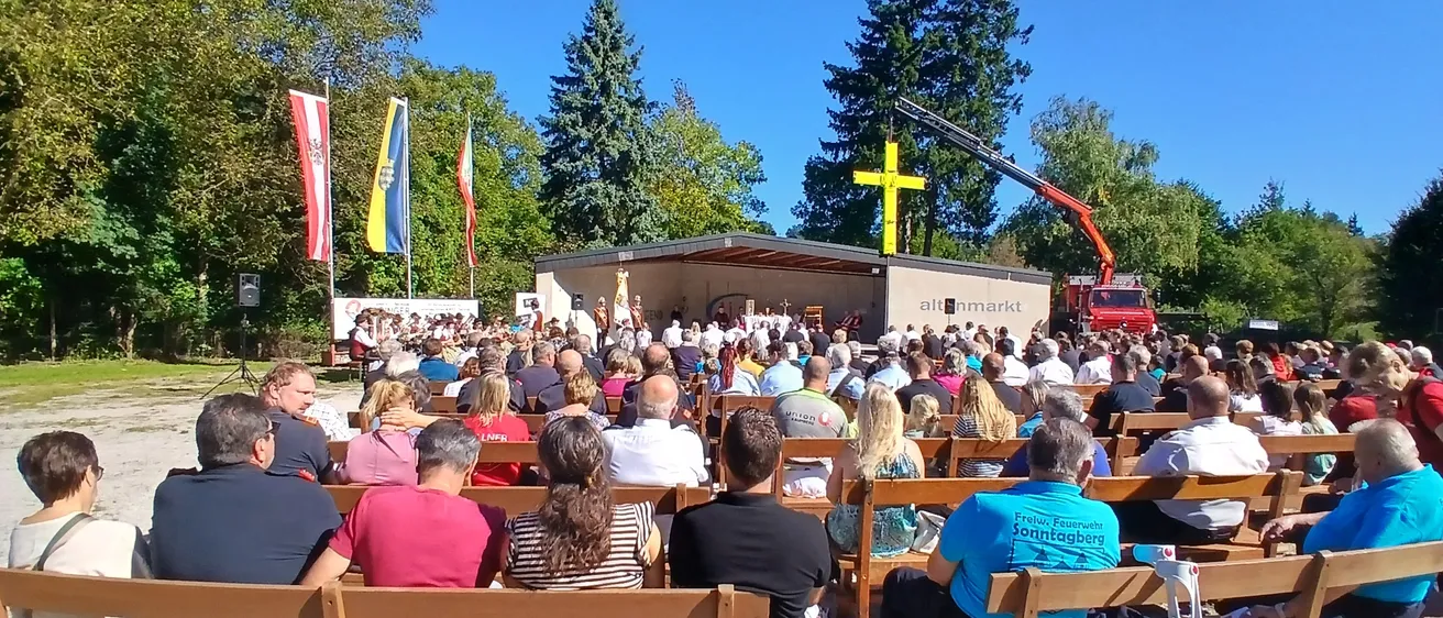 Eine Menschenmenge versammelt sich im Freien für eine Veranstaltung und sitzt auf Holzbänken. Flaggen sind sichtbar und ein Kreuz steht auf einem Gebäude. Ein Baufahrzeug ist in der Nähe geparkt.