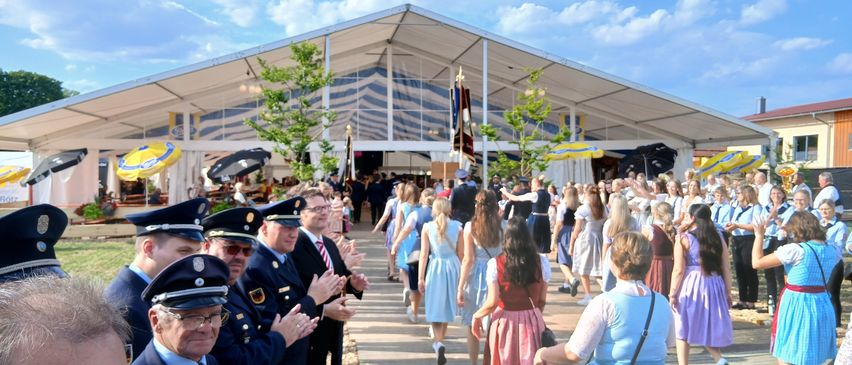 Eine Gruppe von Menschen in blauen Kleidern geht durch einen überdachten Bereich, wobei mehrere Personen in Uniformen klatschen. Der Bereich ist mit weiß-blau gestreiften Wänden, Bäumen und Schirmen umschlossen. Der Himmel darüber ist blau mit weißen Wolken.