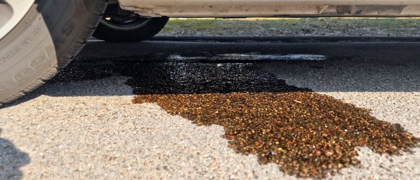 Ein Nahaufnahme des Unterbodens eines Autos, das einen schwarzen Flüssigkeitspool auf der Straße mit Kies in der Nähe zeigt.