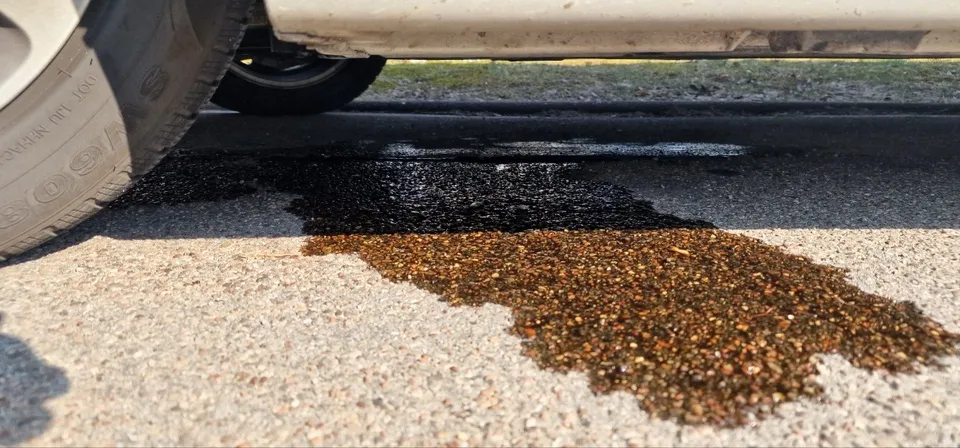 Ein Nahaufnahme des Unterbodens eines Autos, das einen schwarzen Flüssigkeitspool auf der Straße mit Kies in der Nähe zeigt.