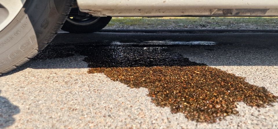Ein Nahaufnahme des Unterbodens eines Autos, das einen schwarzen Flüssigkeitspool auf der Straße mit Kies in der Nähe zeigt.
