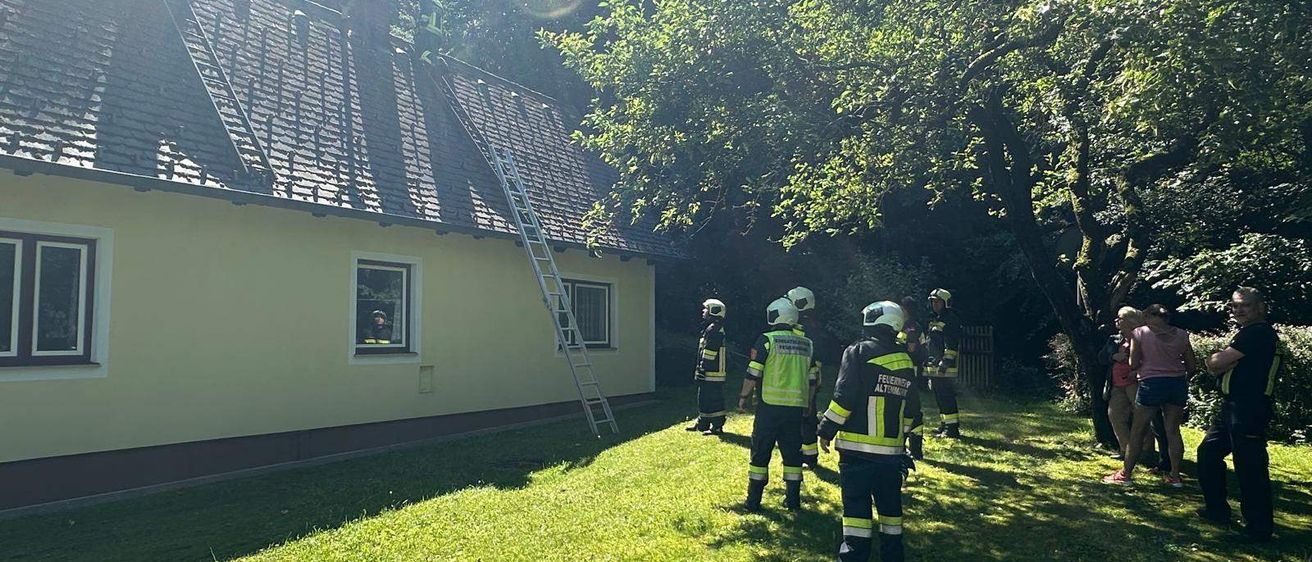 Feuerwehrleute sind auf dem Dach eines Hauses mit einer Leiter. Sie tragen Uniformen. Eine Person ist im Haus und schaut aus dem Fenster.