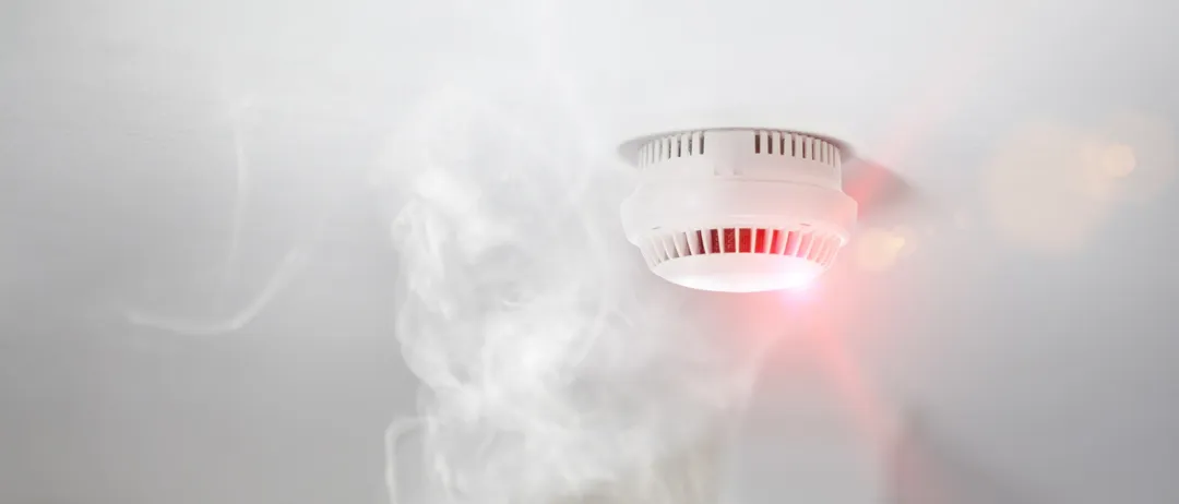 Ein weißer Rauchmelder an der Decke gibt ein rotes Licht ab, was auf einen Alarm hinweist. Rauch steigt von unten auf.