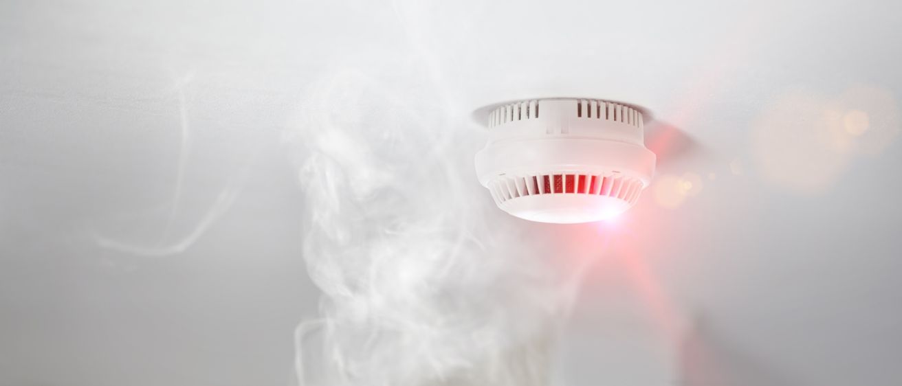 Ein weißer Rauchmelder an der Decke gibt ein rotes Licht ab, was auf einen Alarm hinweist. Rauch steigt von unten auf.