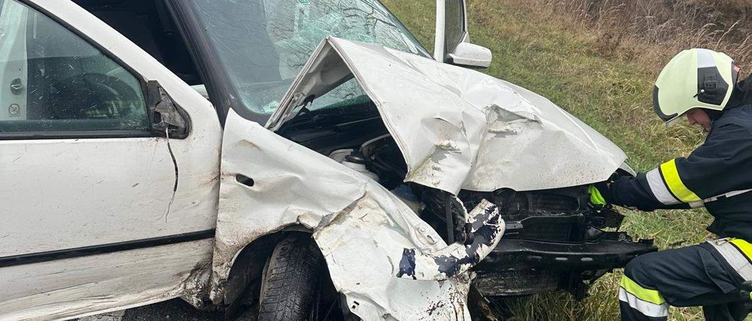 Ein weißer Wagen wurde in einer Kollision schwer beschädigt, wobei die Front stark eingedrückt wurde. Rettungskräfte sind anwesend, darunter ein Feuerwehrmann in Uniform und mit Helm.