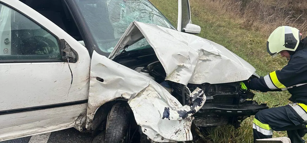 Ein weißer Wagen wurde in einer Kollision schwer beschädigt, wobei die Front stark eingedrückt wurde. Rettungskräfte sind anwesend, darunter ein Feuerwehrmann in Uniform und mit Helm.
