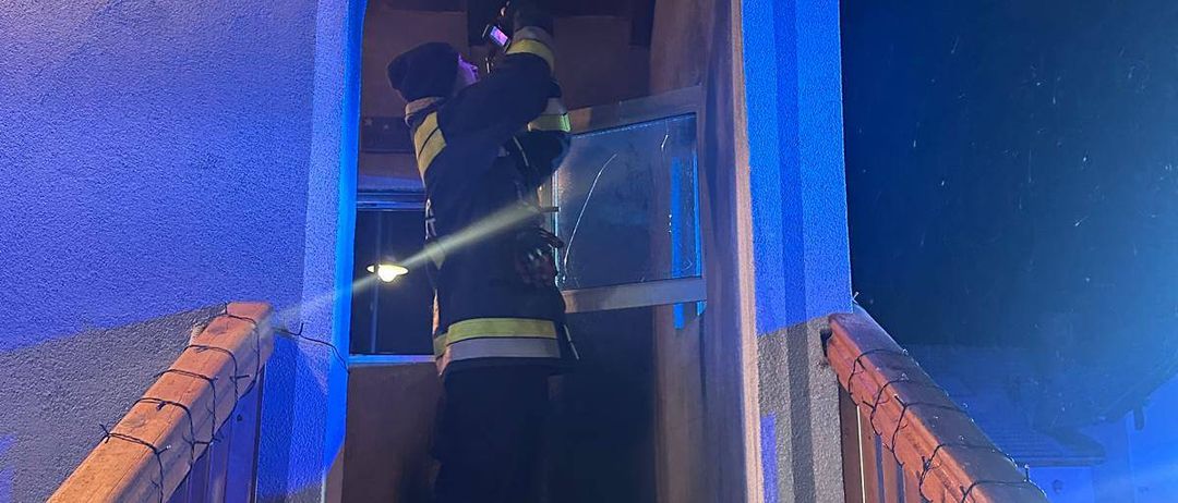 Ein Feuerwehrmann steht nachts in einem Haus, hält eine Taschenlampe in der Hand und schaut auf ein Glasfenster.