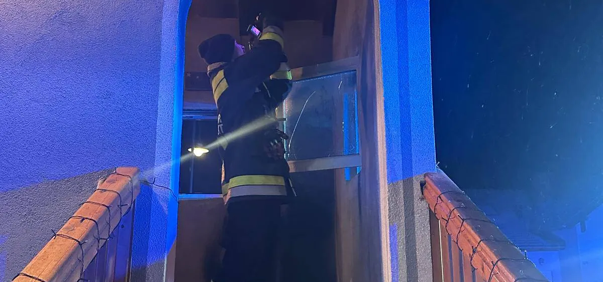 Ein Feuerwehrmann steht nachts in einem Haus, hält eine Taschenlampe in der Hand und schaut auf ein Glasfenster.