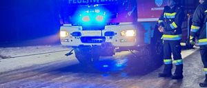 Zwei Feuerwehrleute stehen vor einem Feuerwehrwagen mit blinkenden blauen Lichtern. Der Wagen steht auf einer nassen Straße.
