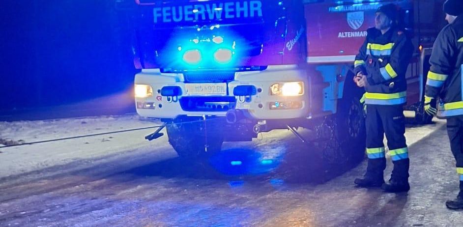 Zwei Feuerwehrleute stehen vor einem Feuerwehrwagen mit blinkenden blauen Lichtern. Der Wagen steht auf einer nassen Straße.