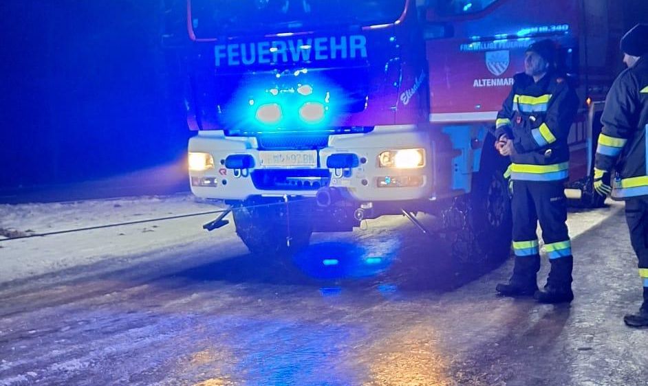 Zwei Feuerwehrleute stehen vor einem Feuerwehrwagen mit blinkenden blauen Lichtern. Der Wagen steht auf einer nassen Straße.