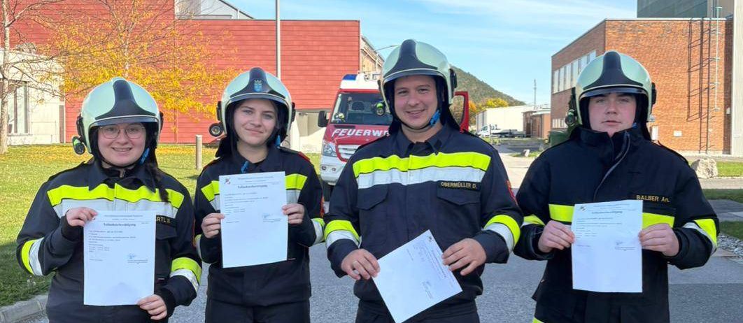 Vier Feuerwehrleute stehen vor einem Gebäude und halten jeweils ein Zertifikat in der Hand. Sie tragen einheitliche Uniformen mit Helmen und Handschuhen. Hinter ihnen ist ein Feuerwehrwagen geparkt.