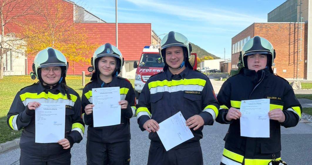 Vier Feuerwehrleute stehen vor einem Gebäude und halten jeweils ein Zertifikat in der Hand. Sie tragen einheitliche Uniformen mit Helmen und Handschuhen. Hinter ihnen ist ein Feuerwehrwagen geparkt.