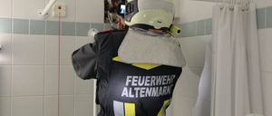 Ein Feuerwehrmann in einem Badezimmer mit einer gebrochenen Wand, der einen Helm und eine reflektierende Weste trägt. Er hält eine Kamera und scheint zu filmen.